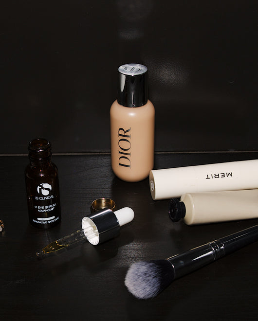 Beauty products on a dark surface, including Dior foundation, eye serum, Merit tubes, and a makeup brush.