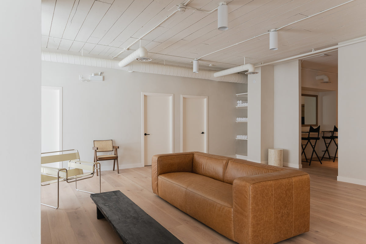 Minimalist living room with a brown leather sofa, modern chairs, white walls, and light wood flooring.