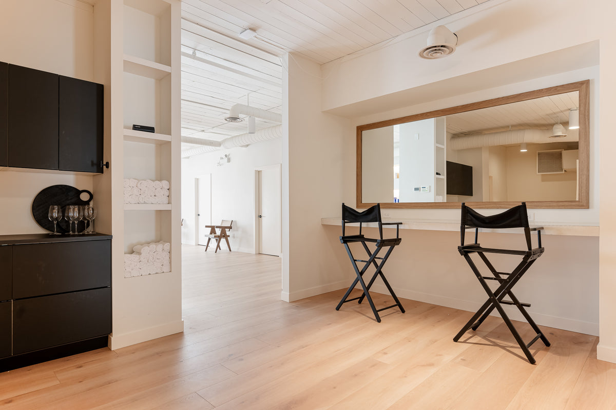 Modern dressing room with a large mirror, two black chairs, shelves with towels, and light wood flooring.