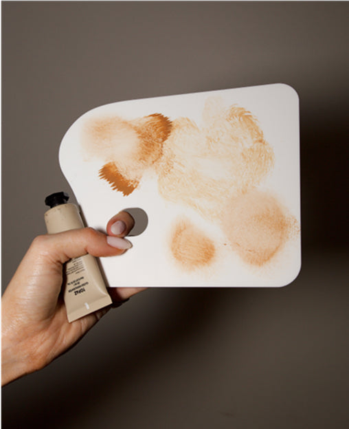 A hand holds a paint tube and palette smeared with various shades of beige and brown makeup.