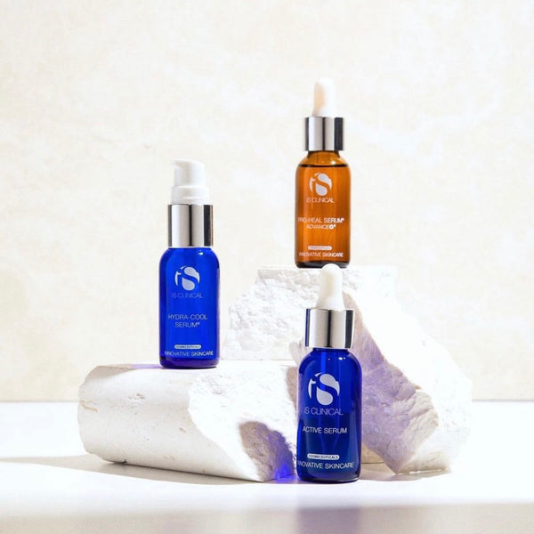Three iS Clinical skincare serum bottles on white stone blocks, two blue and one amber, with dropper and pump lids.