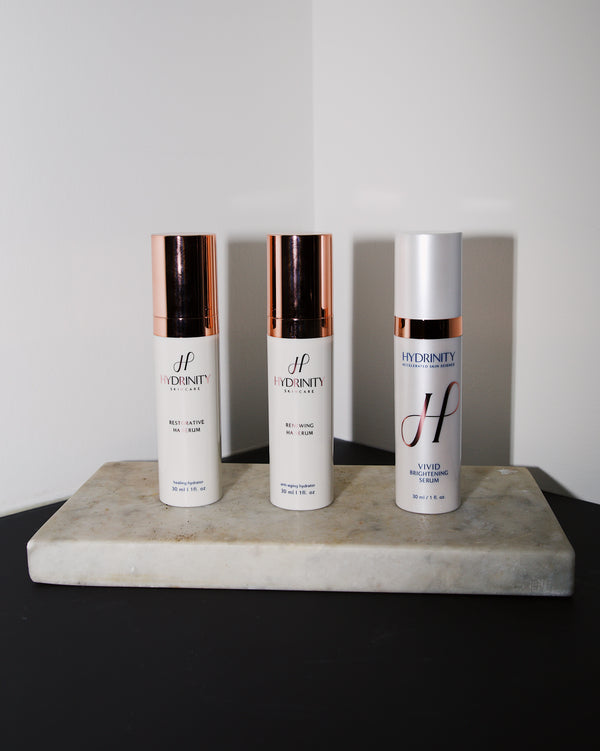 Three Hydrinity skincare bottles with rose gold caps on a rectangular marble tray against a plain background.