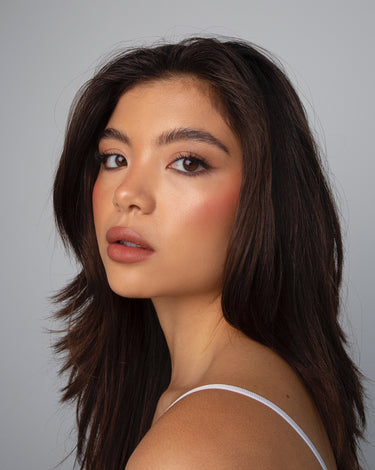 Woman with long dark hair, natural makeup, and a white strap top, looking over her shoulder against a plain background.