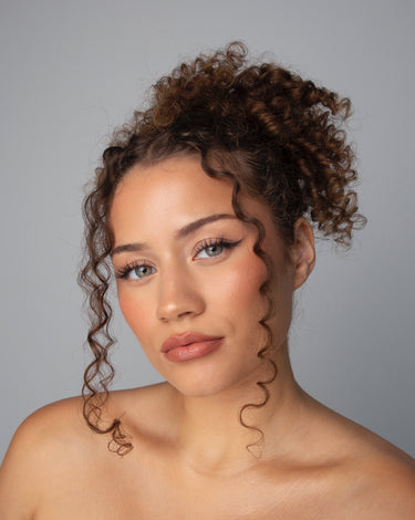 Woman with curly hair and natural makeup poses against a plain gray background, looking directly at the camera.
