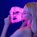 A woman holds the Omnilux Clear LED face mask by Omnilux near her face against a blue background.