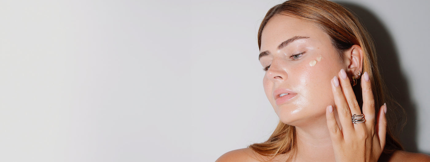 Woman applying cream to her face with her fingers, looking to the side, against a plain light background.