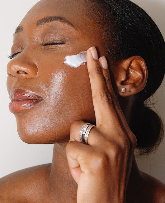 A woman with closed eyes applies a dab of cream to her cheek with her fingers.