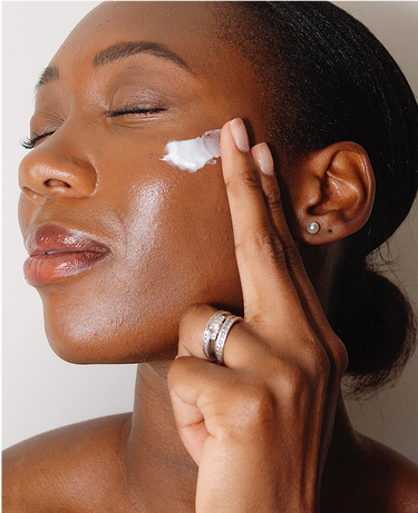 Woman with closed eyes applies moisturizer to her cheek with two fingers, wearing a ring and pearl earring.