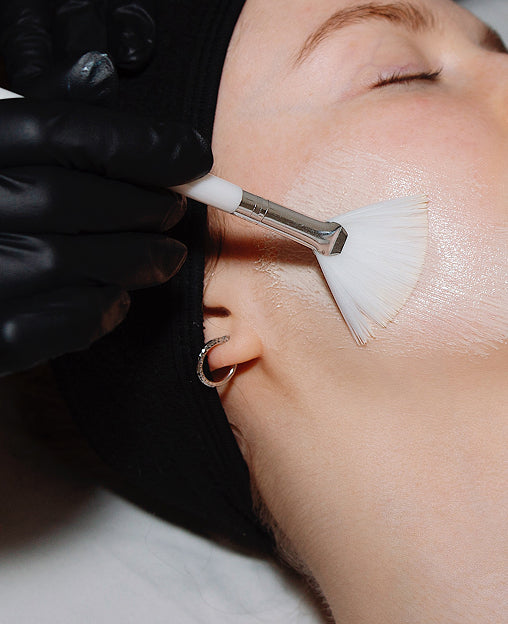 A person receives a facial treatment with a brush applied to their cheek by a gloved hand.