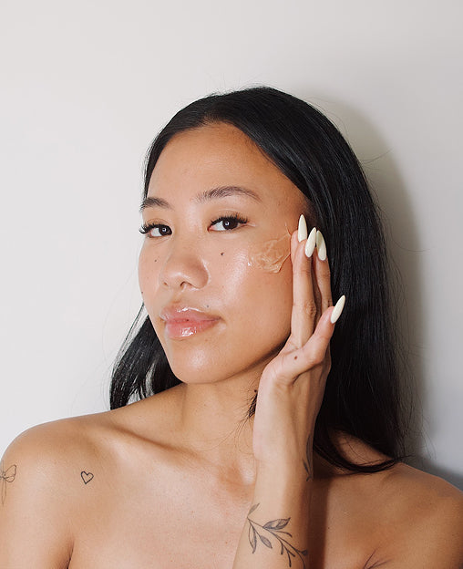 Woman with long black hair applies skincare gel to her cheek, showing glossy skin and decorative tattoos on her arm.