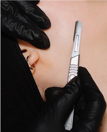 A gloved hand uses a scalpel to perform dermaplaning on a persons cheek near their ear with a hoop earring.