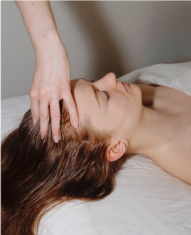 Person lying down with eyes closed, receiving a gentle head massage, covered with a white sheet.