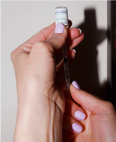 A person with light purple nails draws liquid from a Dysport vial into a syringe.