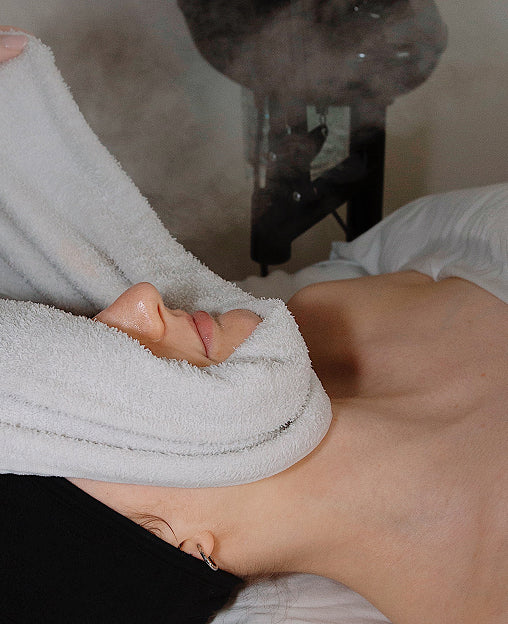 A person receives a facial treatment with a warm towel covering their face while steam is applied.