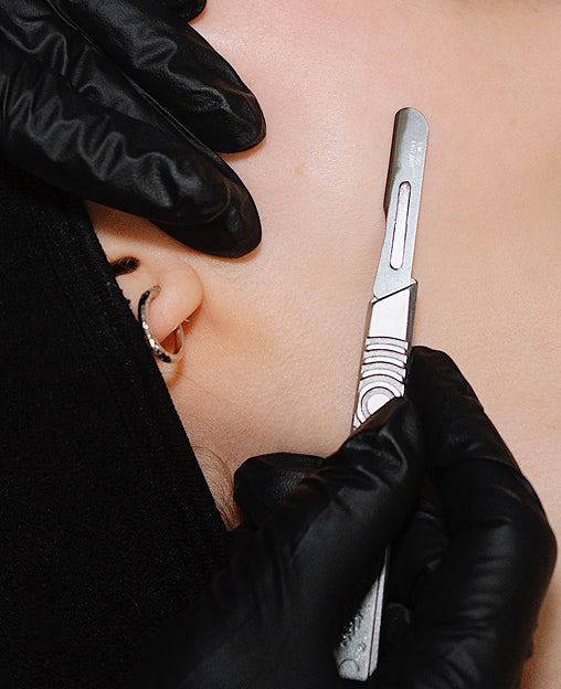 A gloved hand uses a dermaplaning tool on a persons cheek near their ear, which has a hoop earring.
