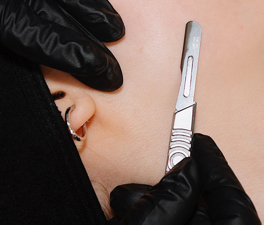 Gloved hands hold a scalpel to a persons cheek, likely performing a skincare or dermaplaning procedure.