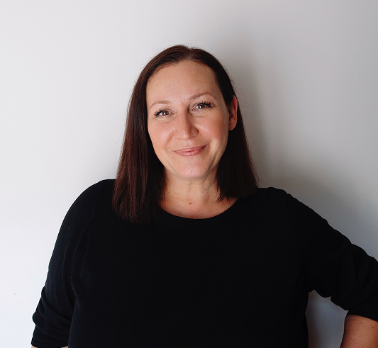 Woman with straight brown hair and a black top smiles at the camera against a plain white background.