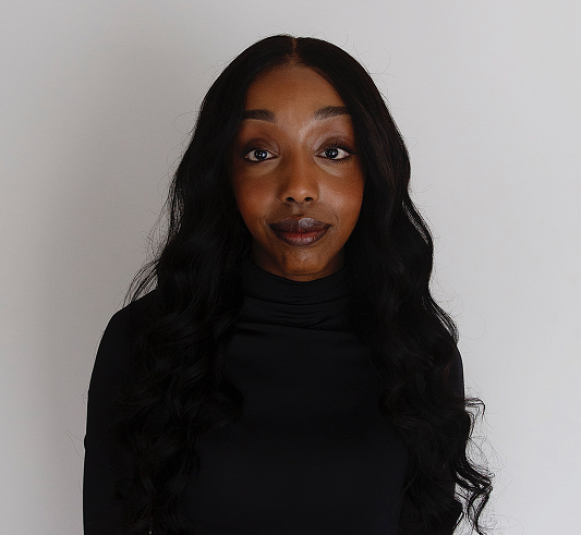 Woman with long wavy black hair, wearing a black turtleneck, standing against a plain white background.