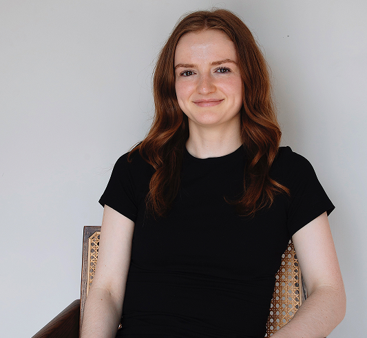 Woman with long red hair, wearing a black shirt, sits on a chair and smiles at the camera against a plain background.