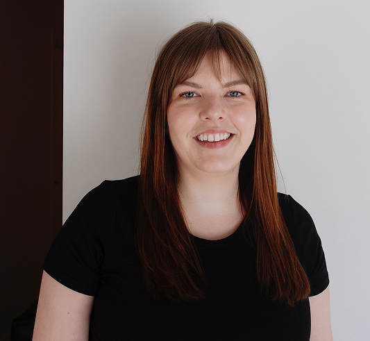 A woman with straight brown hair and a black shirt smiles while standing against a white wall.