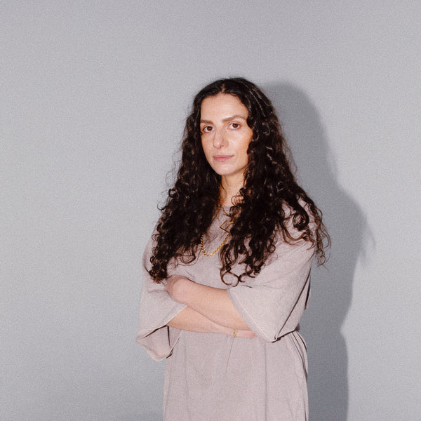 Woman with long curly hair in a light gray dress stands with arms crossed against a plain gray background.