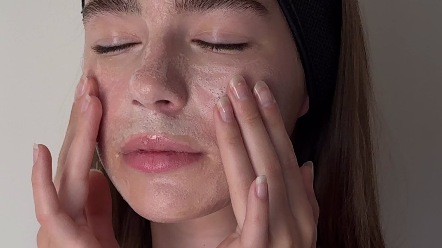 A person with closed eyes gently massages a facial cleanser onto their cheeks with both hands.